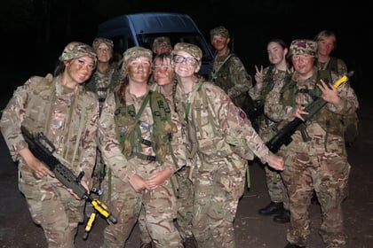 A group of people in military camouflage uniforms and face paint pose together at night, smiling at the camera. Some hold rifles; a van is visible in the background.