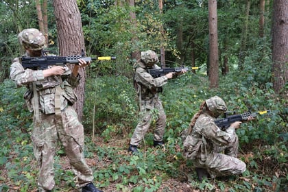 Three people in camouflage military uniforms and helmets aim rifles with yellow tips whilst positioned among trees and dense foliage in a forest.