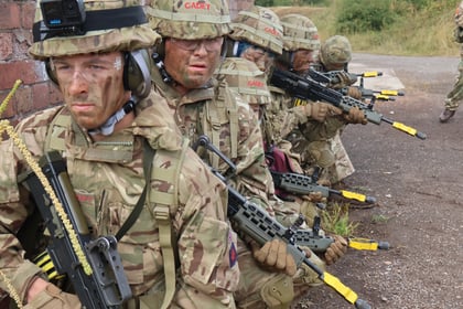 Four soldiers in camouflage gear and helmets crouch in a line beside a brick wall, holding rifles with yellow tips, appearing focused during a training exercise. The scene is outdoors with greenery in the background.