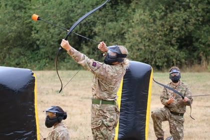 Three people in camouflage uniforms and protective masks participate in an outdoor archery event, aiming foam-tipped arrows whilst kneeling or standing near large black barriers on grassy ground. Trees are visible in the background.