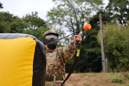 Person in camouflage and face mask aims a bow with a foam-tipped arrow, standing outdoors behind a yellow and black inflatable barrier, with green trees in the background.