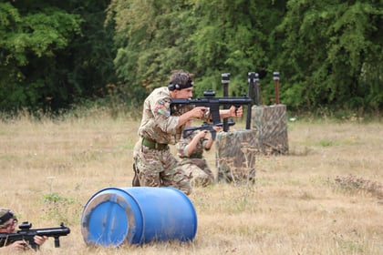 A person in camouflage uniform, holding a large black paintball marker, crouches behind a blue barrel in a grassy field with trees. Another player is partially visible, lying low and aiming their marker.