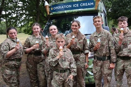 Seven young people in camouflage uniforms stand in front of a camo ice cream van labelled Operation Ice Cream, smiling and holding ice cream cones outdoors on a sunny day.