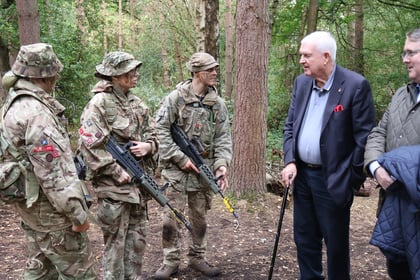 Three soldiers in camouflage uniforms and holding rifles stand in a forest, talking to two men wearing suits. One man uses a walking stick. The group appears to be engaged in conversation among the trees.
