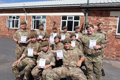 A group of young cadets in camouflage uniforms and berets pose outside a brick building, smiling and holding certificates.