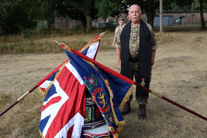 A man in military uniform stands outdoors beside flags and a drum, with another person in uniform in the background on a grassy field.