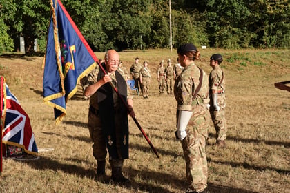 A man in uniform holds a flag and faces a woman in military attire during an outdoor ceremony, with several other soldiers standing in the background on a grassy field.