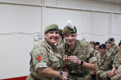 Two people in military uniforms smile and pose for a photo whilst holding an item. Other uniformed individuals are seated and watching in the background inside a plain room.