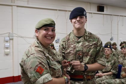 Two people in military uniforms smile at the camera; one is pinning a badge on the other. Both are wearing camouflage and berets. Other uniformed individuals sit in the background.