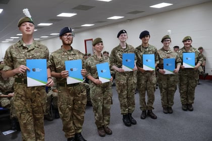 Seven people in military uniforms stand indoors, smiling and holding blue certificates. Other uniformed individuals are seated in the background, suggesting a ceremony or awards event.