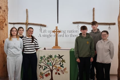 Six young people stand in front of a church altar with a cross, two wooden crosses, and a sword on the wall. A tapestry with a tree design covers the altar. Text on the wall reads, Lift up a living nation...