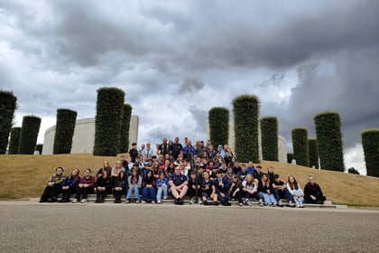 A large group of people sits and stands on outdoor steps in front of tall, trimmed hedges under a cloudy sky.