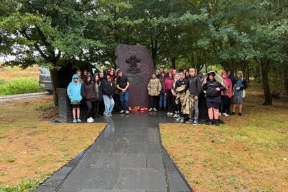 A group of people in hooded raincoats stand around a large stone memorial with wreaths, surrounded by trees on a rainy day.