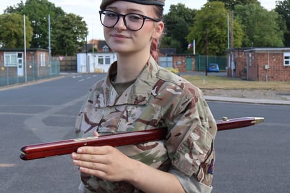 A young person in military camouflage uniform and beret stands outdoors, holding a wooden staff across their chest, with buildings and trees in the background.