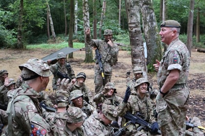 A group of soldiers in camouflage uniforms sit and stand in a forest, listening attentively as a commanding officer speaks. Some soldiers hold rifles; several raise hands or gesture, and tents are faintly visible in the background.