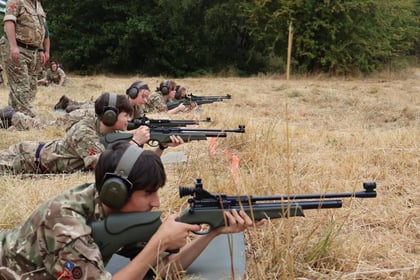 A group of people in camouflage uniforms lies in a row on dry grass, aiming rifles. They wear ear protection and are supervised by an adult in uniform, with trees visible in the background.