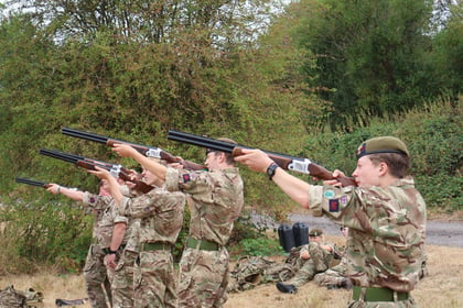Five people in camouflage uniforms stand outdoors, aiming shotguns skywards. More people in similar attire sit or lie on the ground in the background among trees and bushes.