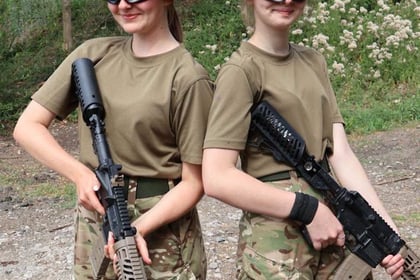 Two people in camouflage trousers, tan shirts, and bucket hats stand back-to-back outdoors, each holding a rifle. They wear protective goggles and smile at the camera. Trees and tall grass are visible in the background.