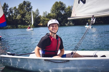 A young child sits on a sailing boat wearing a white helmet and red life jacket
