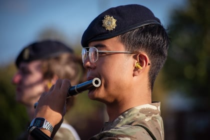 A uniformed person wearing a beret and glasses plays a flute outdoors, with another person in similar attire blurred in the background.