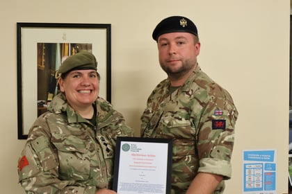 Two people in military uniforms stand indoors, smiling. One holds a framed certificate. There is a framed photo on the wall and an information sign on a table nearby.