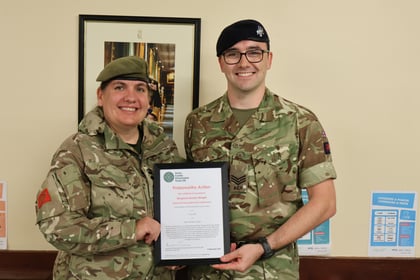 Two people in military uniforms smile whilst holding a framed certificate. They stand indoors in front of beige walls, a framed photo, and posters with text.