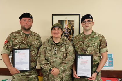 Three people in military uniforms stand indoors, two holding framed certificates. They are smiling,
