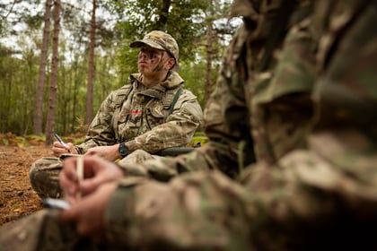 Two cadets in camouflage uniforms and camo cream sit in a forest, writing on notepads.