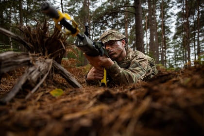 An Army Cadet in camouflage uniform and camo cream lies prone in a forest, aiming a rifle with a yellow attachment.