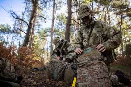 Two Army Cadets in camouflage uniforms and hats kneel in a forest, organising their tactical rucksacks.