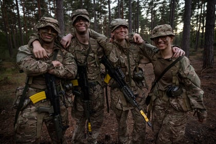 Four Army Cadets in camouflage uniforms with camo cream stand with their arms around each other in a forest, holding rifles.