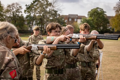 A group of cadets in camouflage uniforms participate in a target shooting exercise with shotguns on an outdoor field.