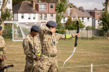A cadet in camouflage uniform aiming a recurve bow on a sports field, whilst another cadet watches nearby.