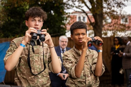 Two young cadets in camouflage uniforms look through binoculars, with an adult in a suit standing blurred in the background.
