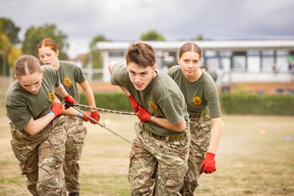 Three cadets in uniform and red gloves pull a rope during an outdoor team activity.