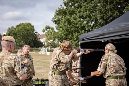 A cadet in a camouflage uniform aims a shotgun at an outdoor shooting range while an instructor watches and another person prepares a firearm nearby.