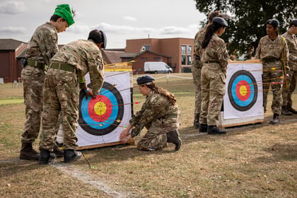 Four cadets in camouflage uniforms retrieve arrows from two archery targets on a grassy field.