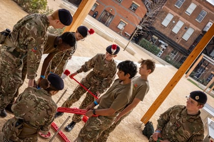 A group of cadets in camouflage uniforms and berets work together to assemble a red plastic frame outdoors.