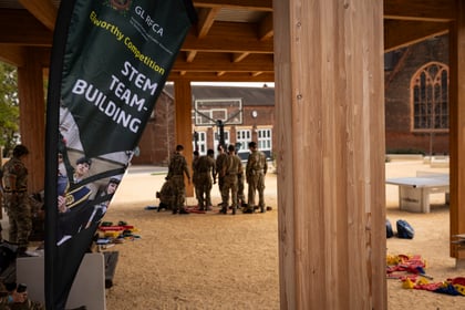 A banner for the Elworthy Competition STEM Team-Building event stands in front of a group of cadets gathered on a sandy outdoor courtyard.
