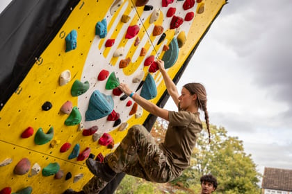 A young woman with a long plait climbs a yellow outdoor bouldering wall covered in colourful holds.