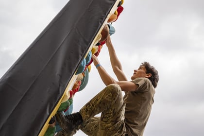 A young man climbs a slanted outdoor rock climbing wall whilst others watch from below.