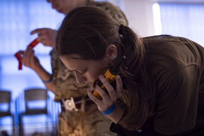 A cadet in uniform writes on a piece of paper while holding a portable radio to their ear, as another person stands in the background.