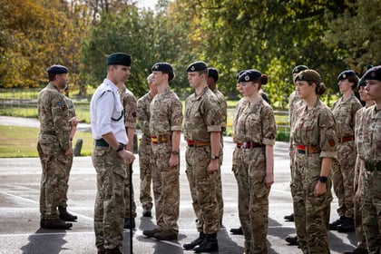 A military officer in a white shirt and camouflage trousers inspects a row of Army Cadets standing at attention in camouflage uniforms.