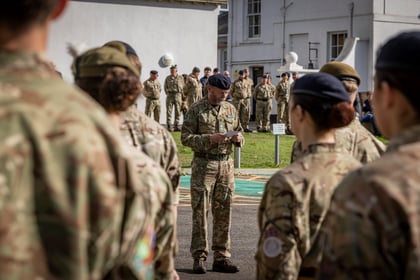 An officer in camouflage uniform reads from a paper whilst standing before a group of Army Cadets in formation.