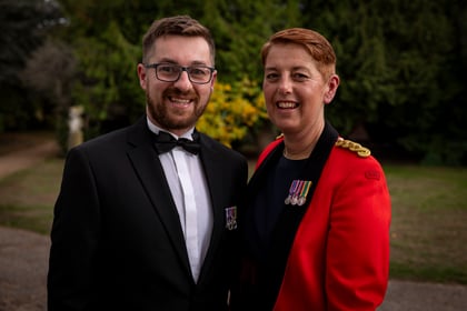 A man in a black dinner jacket and a woman in a red ceremonial military jacket with medals stand together outdoors.