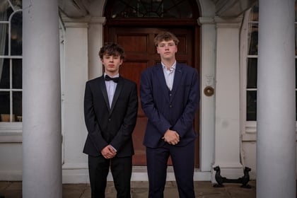 Two young men wear suits and pose for a photo in front of a wooden door