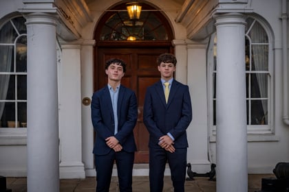 Two young men in suits standing in front of a doorway flanked by white columns.