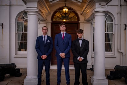 Three young men in formal suits stand in a row under the arched portico of a white building.