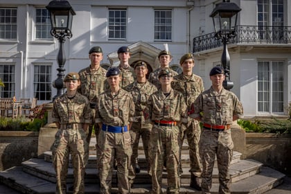 A group of nine Army Cadets in camouflage uniforms pose for a photo on the steps of a white building.