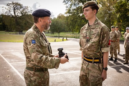 A military officer in camouflage uniform presents a small boot-shaped trophy to a young soldier standing to attention.